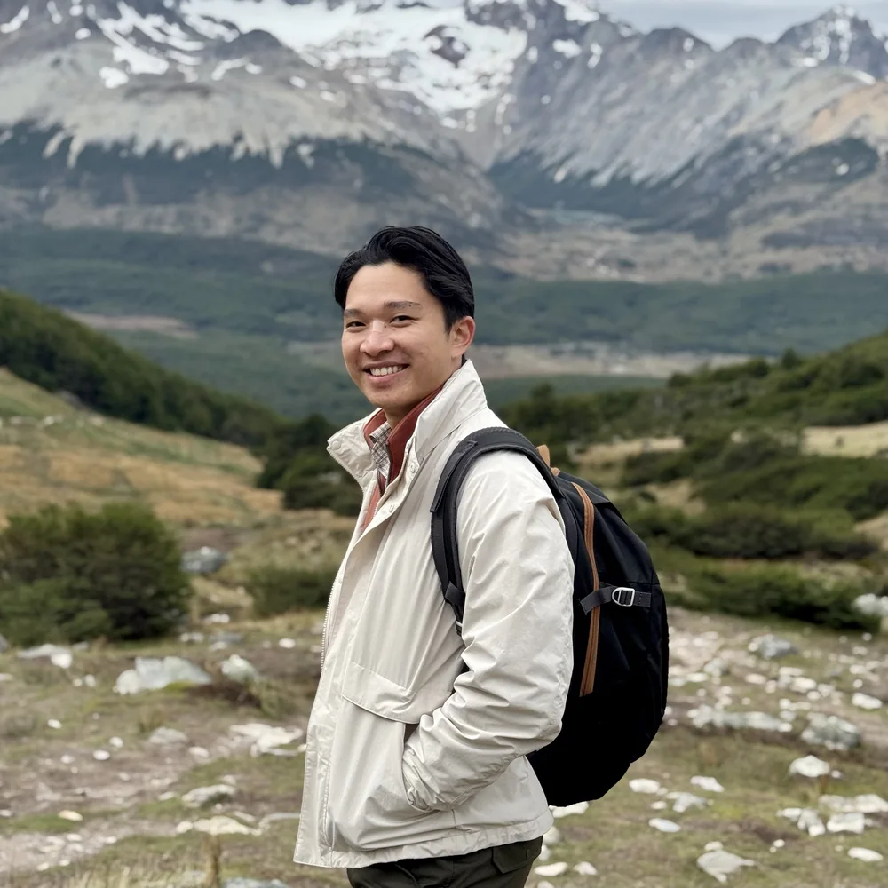Rio smiling on a mountain trail, wearing a cream jacket and a backpack